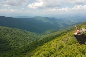 girl sitting on edge of a cliff, hands in the air, mountain view, green, day hike essentials